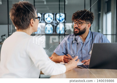 Male healthcare professional in blue scrubs discusses medical results with a female patient in a modern clinic setting with medical imaging displayed in the background Male healthcare professional in blue scrubs discusses medical results with a female patient in a modern clinic setting with medical imaging displayed in the background 140039866