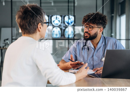 Male healthcare professional in blue scrubs discusses medical results with female patient in modern clinic, medical imaging displayed on wall behind them Male healthcare professional in blue scrubs discusses medical results with female patient in modern clinic, medical imaging displayed on wall behind them 140039867