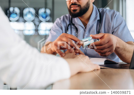 Male healthcare professional in blue scrubs uses a pulse oximeter on a patient's finger during a consultation in a modern medical office with medical equipment in the background Male healthcare professional in blue scrubs uses a pulse oximeter on a patient's finger during a consultation in a modern medical office with medical equipment in the background 140039871
