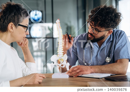 Male healthcare professional in blue scrubs explaining spinal model to female patient with short gray hair in a modern medical office setting Male healthcare professional in blue scrubs explaining spinal model to female patient with short gray hair in a modern medical office setting 140039876