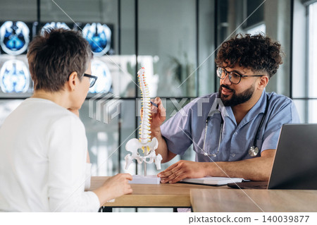 Male healthcare professional explaining spinal anatomy to female patient using a model in a modern medical office with X-ray images in the background 140039877