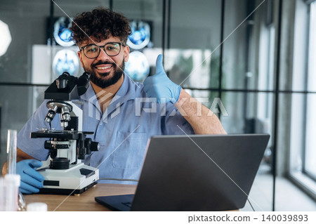 Male laboratory technician in blue shirt and gloves gives a thumbs-up gesture beside a microscope and laptop on a wooden table in a modern lab setting Male laboratory technician in blue shirt and gloves gives a thumbs-up gesture beside a microscope and laptop on a wooden table in a modern lab setting 140039893