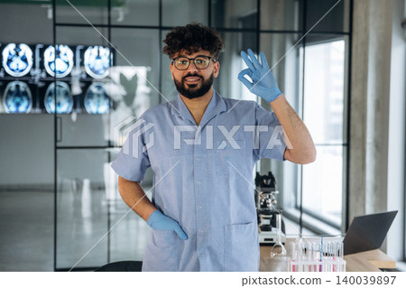 Male healthcare professional in blue scrubs gestures with hand raised while standing in a modern laboratory with medical equipment and glass walls in the background Male healthcare professional in blue scrubs gestures with hand raised while standing in a modern laboratory with medical equipment and glass walls in the background 140039897