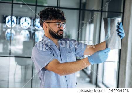 Male healthcare professional in blue scrubs examines X-ray image while standing in a modern medical office with brain scans displayed in the background Male healthcare professional in blue scrubs examines X-ray image while standing in a modern medical office with brain scans displayed in the background 140039908
