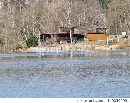 Rustic house beside tranquil lake. Calm lakeside setting featuring bare birch and reflective waters 140039999