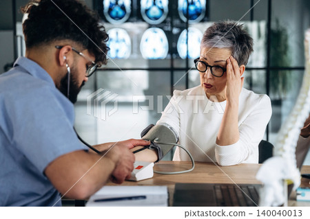 Male healthcare professional measuring blood pressure of a female patient in a modern clinic with medical charts and anatomical models visible in the background Male healthcare professional measuring blood pressure of a female patient in a modern clinic with medical charts and anatomical models visible in the background 140040013