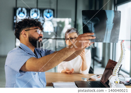 Male healthcare professional in blue shirt examines knee X-ray while discussing with female patient in modern medical office with spine model and medical images in background 140040021