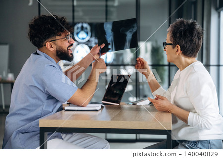 Male healthcare professional in blue scrubs discusses X-ray results with female patient in modern clinic setting, laptop and medical images visible on table Male healthcare professional in blue scrubs discusses X-ray results with female patient in modern clinic setting, laptop and medical images visible on table 140040029
