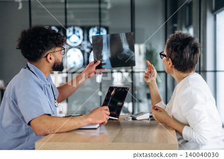 Male healthcare professional in blue scrubs discusses X-ray results with female doctor in white coat, modern medical office with large windows and equipment visible Male healthcare professional in blue scrubs discusses X-ray results with female doctor in white coat, modern medical office with large windows and equipment visible 140040030