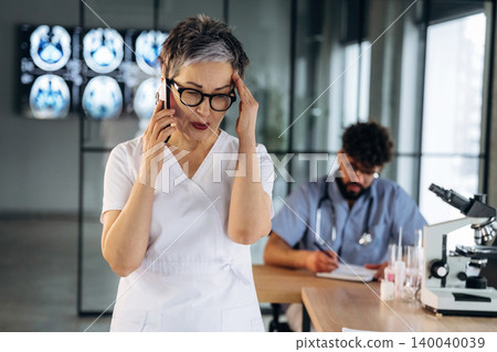 Female healthcare professional in white scrubs holds her head while talking on the phone, with a male colleague and medical equipment in a modern clinic setting Female healthcare professional in white scrubs holds her head while talking on the phone, with a male colleague and medical equipment in a modern clinic setting 140040039