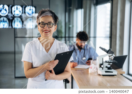Female healthcare professional in white uniform holds clipboard, smiling in modern medical office with male colleague working at desk and microscope in background Female healthcare professional in white uniform holds clipboard, smiling in modern medical office with male colleague working at desk and microscope in background 140040047