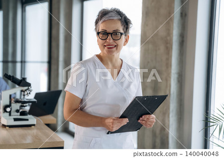 Middle-aged female healthcare professional in white scrubs holds a clipboard while standing in a modern laboratory with microscopes and large windows in the background 140040048