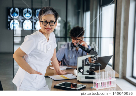 Female healthcare professional in white scrubs smiles confidently while standing beside a microscope and test tubes in a modern medical office with a colleague in the background 140040056