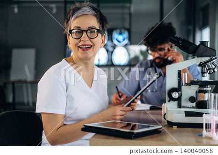 Female healthcare professional in white scrubs smiles while holding a tablet, with a male colleague examining samples at a microscope in a modern laboratory setting Female healthcare professional in white scrubs smiles while holding a tablet, with a male colleague examining samples at a microscope in a modern laboratory setting 140040058