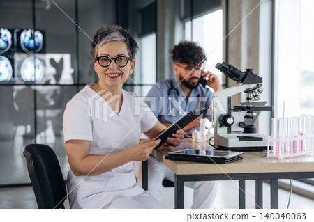 Female healthcare professional in white scrubs holding a clipboard, seated at a desk with a microscope and laboratory equipment in a modern medical office setting Female healthcare professional in white scrubs holding a clipboard, seated at a desk with a microscope and laboratory equipment in a modern medical office setting 140040063