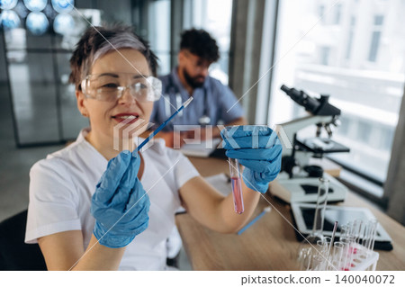 Female laboratory technician in white coat holds test tube with pink liquid and pipette, with microscope and male colleague in background inside modern lab 140040072