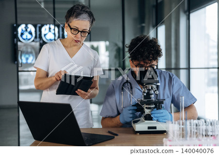Female healthcare professional observes male colleague using microscope in laboratory, with digital tablet and laptop on table, medical images displayed in background Female healthcare professional observes male colleague using microscope in laboratory, with digital tablet and laptop on table, medical images displayed in background 140040076