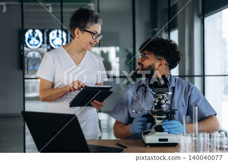 Female healthcare professional in white scrubs discusses findings with male colleague in blue scrubs, laboratory setting with microscope and medical imaging in background 140040077