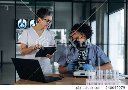 Healthcare professionals discussing laboratory results in a modern medical office with a microscope, laptop, and test tubes on the table 140040079