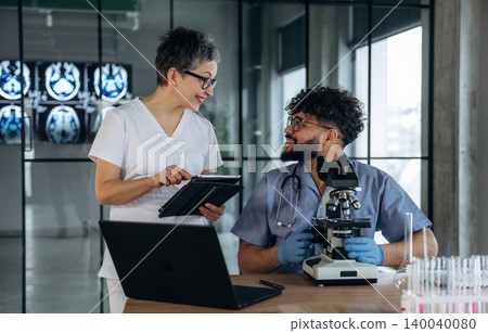 Female healthcare professional in white scrubs discusses findings with male colleague in blue scrubs, laboratory setting with microscope and medical imaging in background Female healthcare professional in white scrubs discusses findings with male colleague in blue scrubs, laboratory setting with microscope and medical imaging in background 140040080