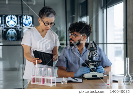 Female healthcare professional in white scrubs discusses findings with male colleague in blue scrubs, laboratory setting with microscope and medical imaging in background Female healthcare professional in white scrubs discusses findings with male colleague in blue scrubs, laboratory setting with microscope and medical imaging in background 140040085