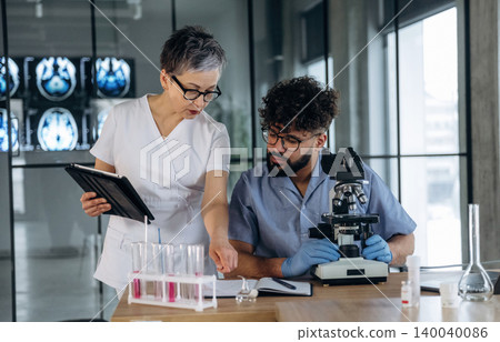 Female healthcare professional in scrubs discusses laboratory results with male colleague at a microscope, test tubes with colored liquids on a table in a modern lab setting 140040086