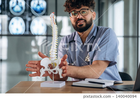 Male healthcare professional in blue scrubs demonstrates a spinal model on a wooden table with medical imaging displayed in the background 140040094