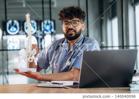 Male healthcare professional in scrubs holds a spinal model while sitting at a desk with a laptop and medical images displayed in the background Male healthcare professional in scrubs holds a spinal model while sitting at a desk with a laptop and medical images displayed in the background 140040096