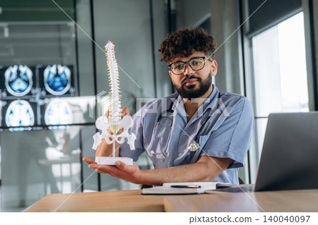 Male healthcare professional in scrubs holding a spinal model while seated at a desk with medical imaging displayed in the background Male healthcare professional in scrubs holding a spinal model while seated at a desk with medical imaging displayed in the background 140040097