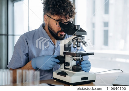 Male scientist wearing blue lab coat and gloves examines samples under a microscope in a bright laboratory with glass test tubes on the table Male scientist wearing blue lab coat and gloves examines samples under a microscope in a bright laboratory with glass test tubes on the table 140040100