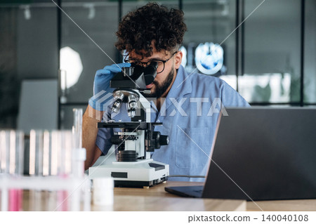 Male laboratory technician wearing blue shirt and gloves examines samples under a microscope in a modern lab setting with a laptop and test tubes on the table Male laboratory technician wearing blue shirt and gloves examines samples under a microscope in a modern lab setting with a laptop and test tubes on the table 140040108