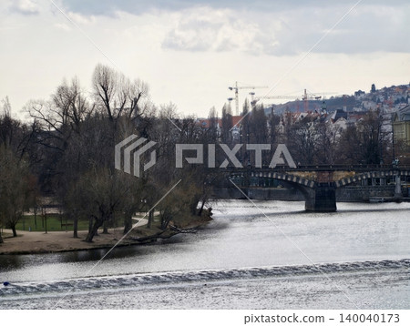 Urban river scenery. Industrial skyline with flowing water. Distant cranes and leafless trees over water Urban river scenery. Industrial skyline with flowing water. Distant cranes and leafless trees over water 140040173