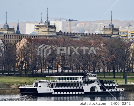 Passenger ferry docked beside treelined bank. Calm reflective water with cityscape in background Passenger ferry docked beside treelined bank. Calm reflective water with cityscape in background 140040241