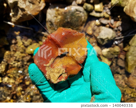 Gloved hand holds a beautiful multi-colored piece of raw flint, showcasing its rugged texture and natural hues against a blurred rocky stream background Gloved hand holds a beautiful multi-colored piece of raw flint, showcasing its rugged texture and natural hues against a blurred rocky stream background 140040338