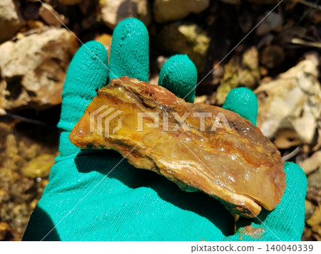 Gloved hand gripping a raw, wet chalcedony chunk, its honeyed and russet tones glinting under the sun. A fresh geological find from a natural, rocky stream setting Gloved hand gripping a raw, wet chalcedony chunk, its honeyed and russet tones glinting under the sun. A fresh geological find from a natural, rocky stream setting 140040339
