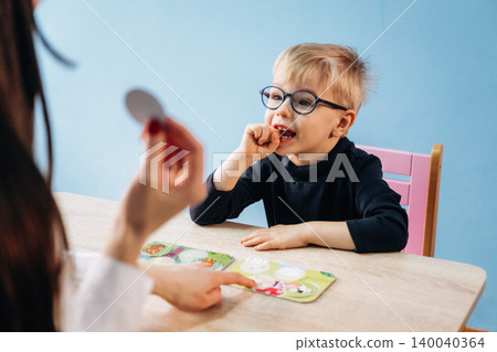 Preschool child with glasses engaged in learning activity with teacher, colorful educational cards displayed on table in bright classroom setting Preschool child with glasses engaged in learning activity with teacher, colorful educational cards displayed on table in bright classroom setting 140040364