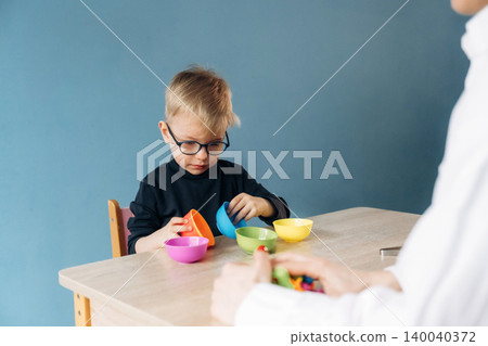 Preschool child with glasses engaged in developmental play with colorful bowls at a wooden table, teacher's hands visible in the foreground, blue wall background Preschool child with glasses engaged in developmental play with colorful bowls at a wooden table, teacher's hands visible in the foreground, blue wall background 140040372
