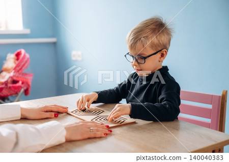 Preschool child with glasses engages in a maze activity with teacher's hand visible, sitting at a wooden table in a bright classroom with blue walls Preschool child with glasses engages in a maze activity with teacher's hand visible, sitting at a wooden table in a bright classroom with blue walls 140040383