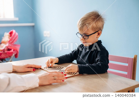 Preschool child with glasses engages in a developmental activity with a teacher's hand visible, colorful classroom setting with blue walls and wooden table Preschool child with glasses engages in a developmental activity with a teacher's hand visible, colorful classroom setting with blue walls and wooden table 140040384