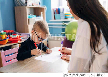 Preschool child with glasses using red marker while drawing on paper with teacher assisting at a colorful classroom table filled with educational toys and materials 140040388