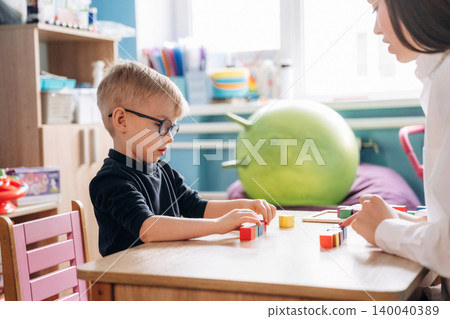 Preschool child with glasses engaged in developmental activities with teacher at a wooden table in a colorful classroom filled with educational materials and toys Preschool child with glasses engaged in developmental activities with teacher at a wooden table in a colorful classroom filled with educational materials and toys 140040389