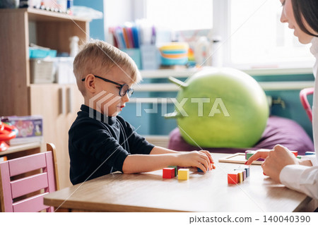 Preschool child with glasses engaged in developmental activities at a wooden table with colorful blocks, while a teacher assists in a bright classroom setting Preschool child with glasses engaged in developmental activities at a wooden table with colorful blocks, while a teacher assists in a bright classroom setting 140040390