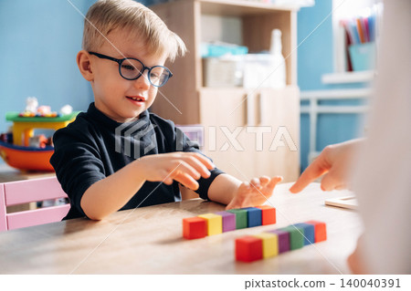 Preschool child with glasses engaged in developmental activity with colorful building blocks at a wooden table in a bright classroom setting Preschool child with glasses engaged in developmental activity with colorful building blocks at a wooden table in a bright classroom setting 140040391