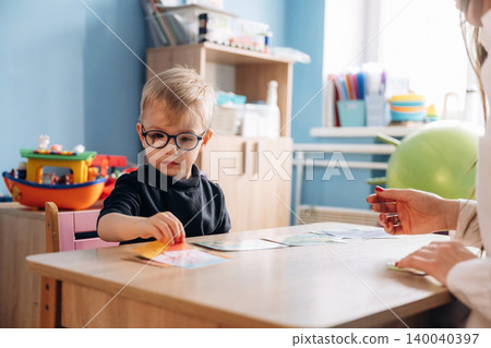 Preschool child with glasses engaged in developmental activity at a wooden table with a teacher's hand visible, colorful toys and educational materials in a bright classroom Preschool child with glasses engaged in developmental activity at a wooden table with a teacher's hand visible, colorful toys and educational materials in a bright classroom 140040397