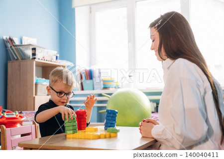 Preschool child with glasses stacking colorful blocks while teacher observes attentively in a bright classroom with educational materials and toys visible in the background Preschool child with glasses stacking colorful blocks while teacher observes attentively in a bright classroom with educational materials and toys visible in the background 140040411