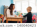 Preschool teacher interacts with young child at a wooden table in a colorful classroom filled with educational toys and materials during a developmental activity 140040423