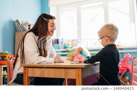 Preschool teacher interacts with young child at a wooden table in a colorful classroom filled with educational toys and materials during a developmental activity 140040423