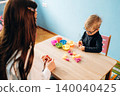 Preschool child with teacher engaged in developmental play at a wooden table, colorful bowls filled with materials visible in a bright classroom setting 140040425