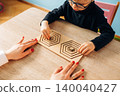 Preschool child with glasses engages in hands-on learning with a teacher's guidance, manipulating geometric wooden shapes on a light wooden table in a colorful classroom 140040427