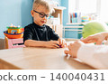 Preschool child with glasses engaged in developmental play with teacher at a wooden table, colorful toys and educational materials visible in a bright classroom setting 140040431